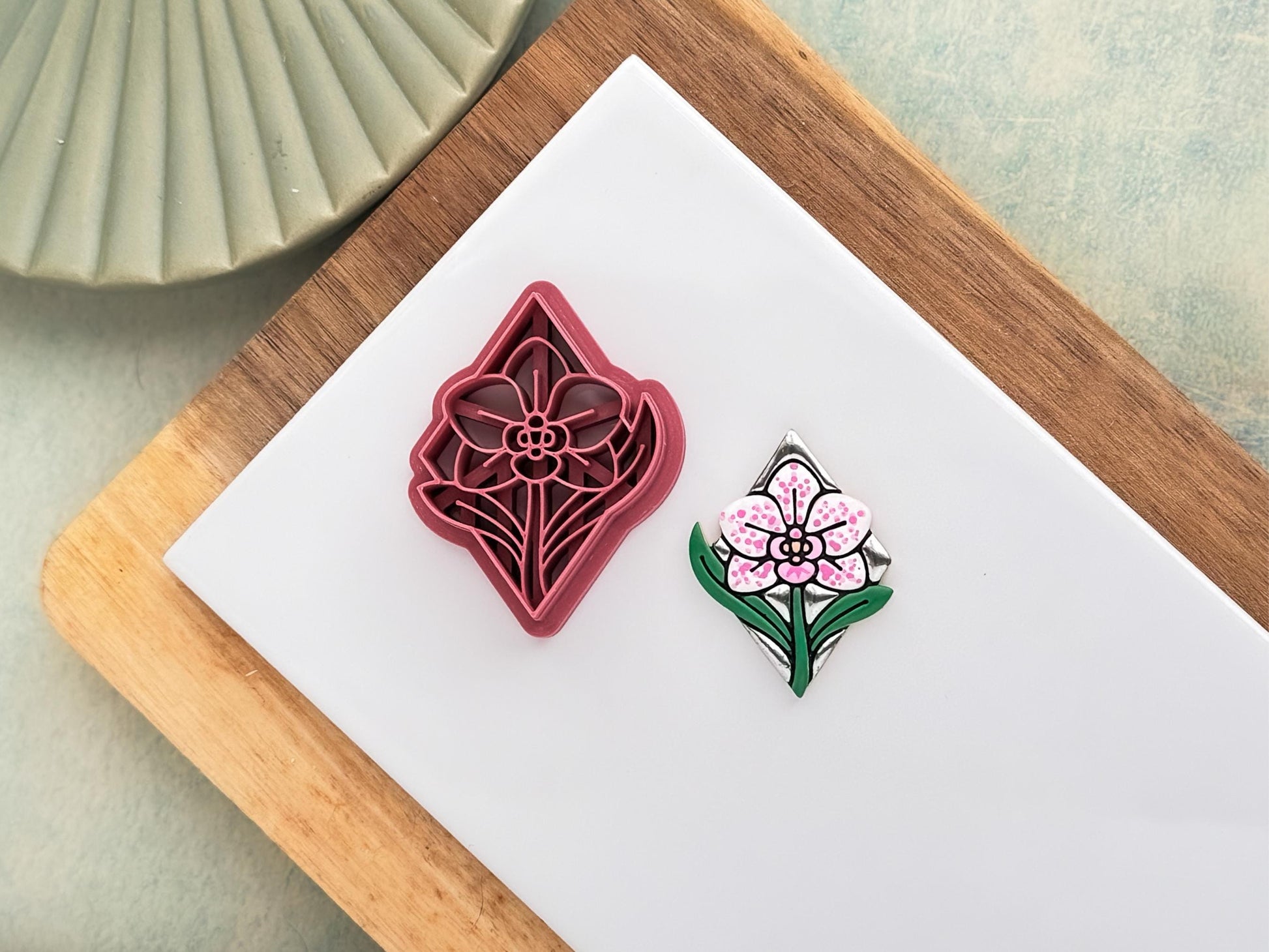 Pink clay cutter in the shape of a diamond with orchid flower and leaf stamp details. Next to it is a polymer clay sample cut out painted in chrome silver, green, white, and pink.