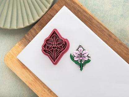 Pink clay cutter in the shape of a diamond with orchid flower and leaf stamp details. Next to it is a polymer clay sample cut out painted in chrome silver, green, white, and pink.