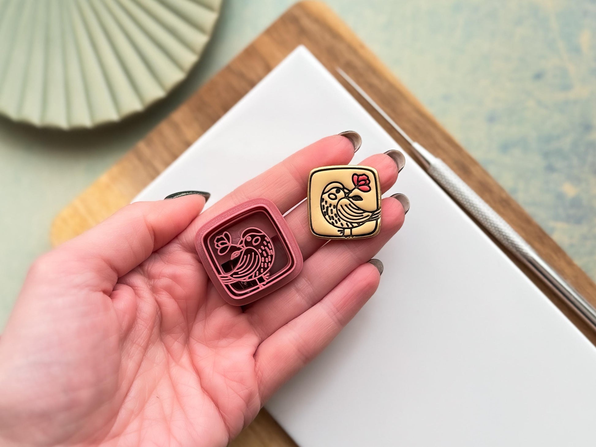Small square clay cutters with a cute bird holding a flower stamp detail. Next to it is a beige polymer clay cut out of the smaller cutter. Both are being held in a hand for scale