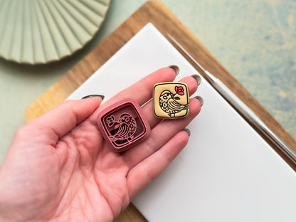 Small square clay cutters with a cute bird holding a flower stamp detail. Next to it is a beige polymer clay cut out of the smaller cutter. Both are being held in a hand for scale