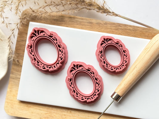Three vintage frame polymer clay cutters with swirling gilded and ornate patterns in different sizes on a white clay baking tile.