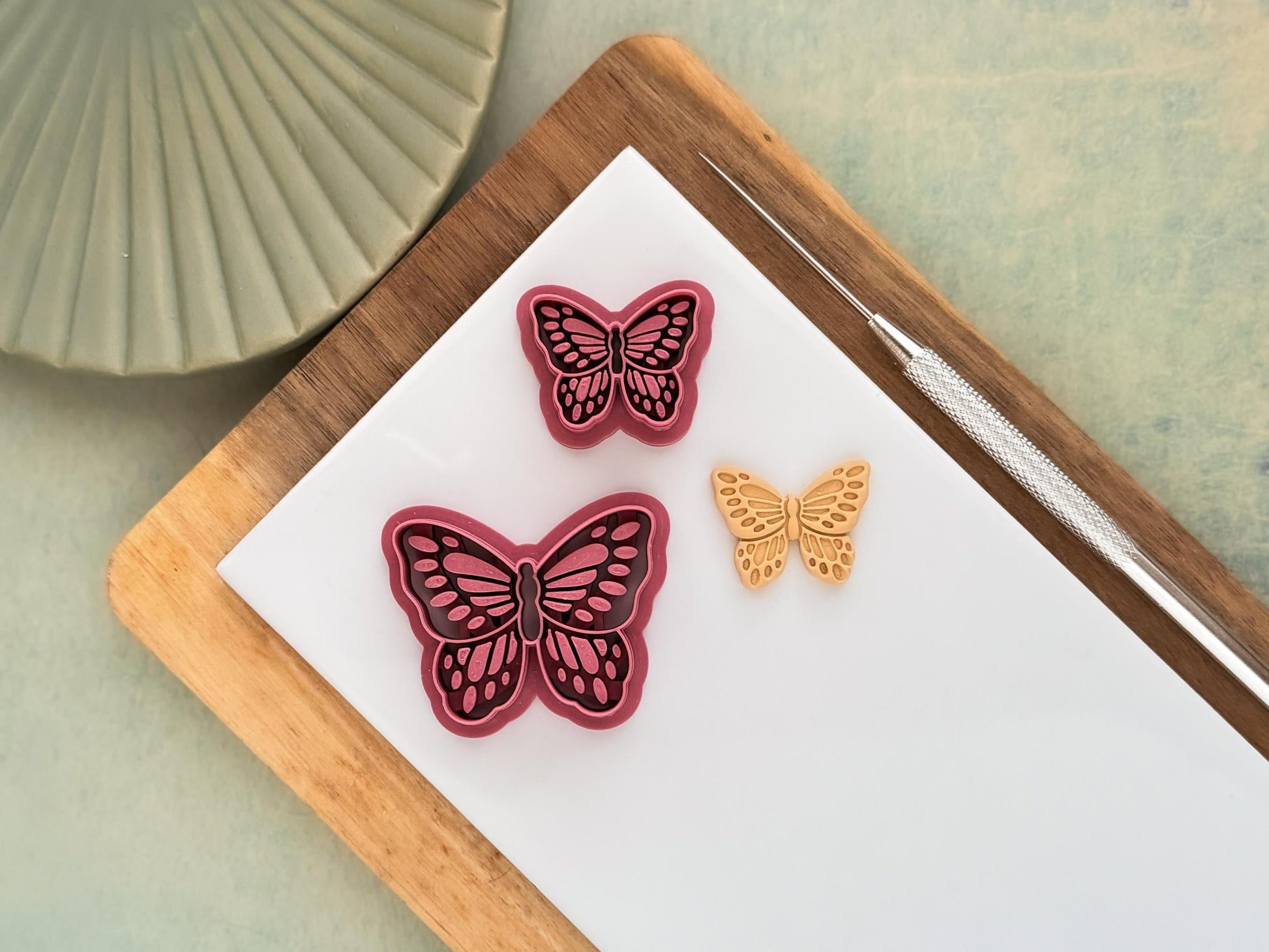 Two pink detailed butterfly clay cutters with embossing details. Next to the cutters is a beige polymer clay sample made with the cutters. These items are on a white baking tile.