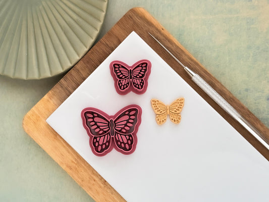 Two pink detailed butterfly clay cutters with embossing details. Next to the cutters is a beige polymer clay sample made with the cutters. These items are on a white baking tile.