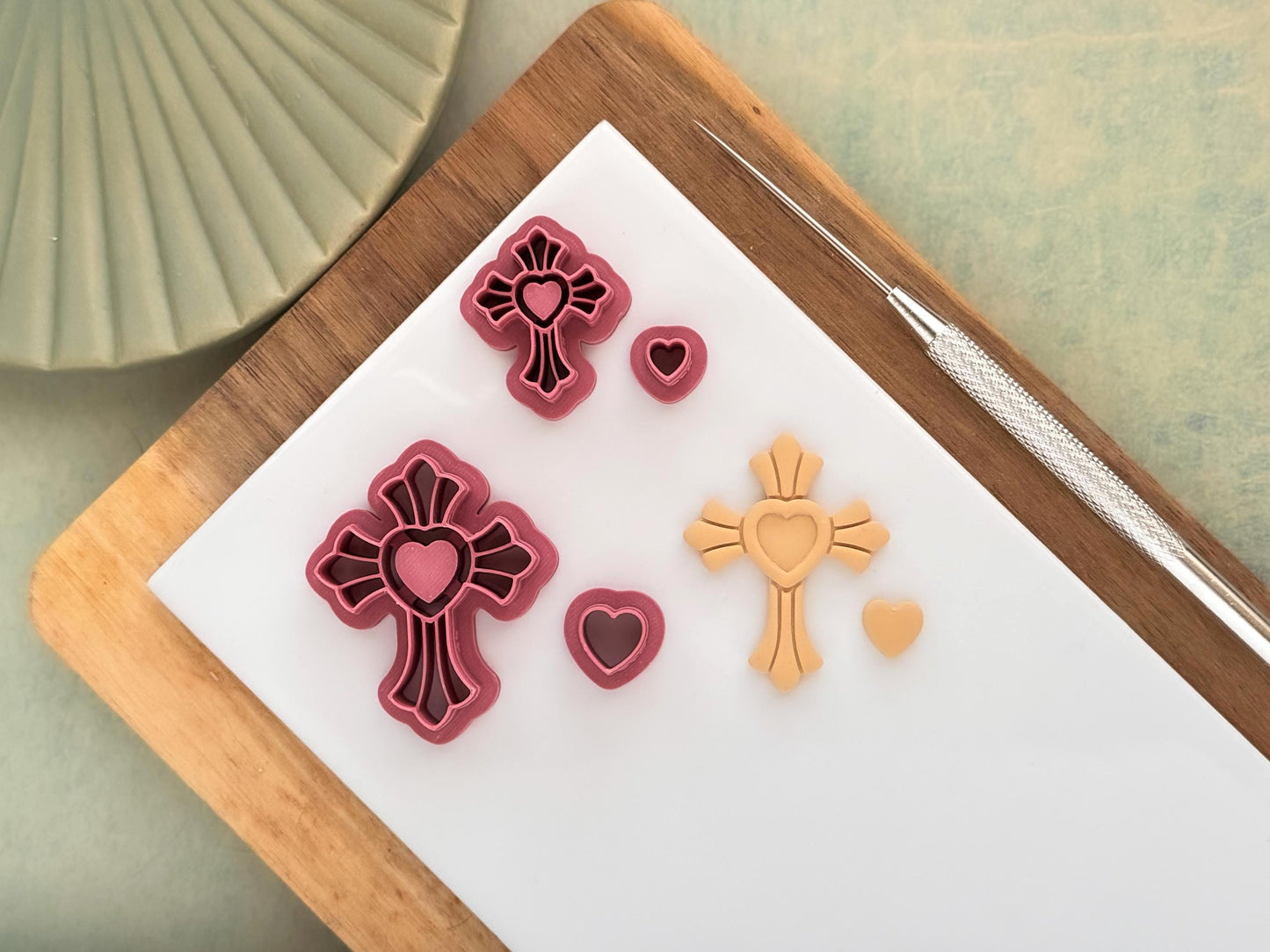 This image shows two dark pink bezel style clay cutters in the shape of a cross and separate heart cutter. Next to the cutters is a polymer clay cut out.