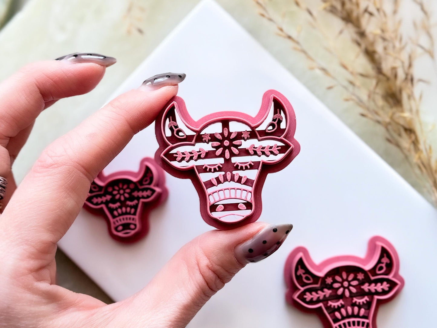 A pink cow-shaped clay cutter with a detailed floral design. The cutter is held in a hand, with a second cutter visible nearby. The design includes intricate cutouts, creating a decorative effect.