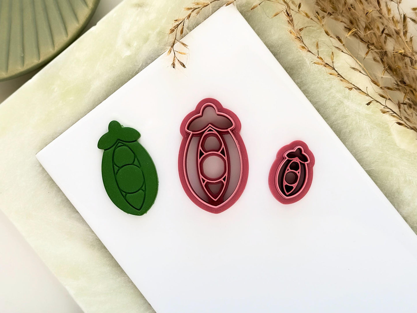 Pea pod shaped clay cutter. Vegetable themed craft cutter with pea stamp details in two sizes, one small for making stud earrings and the other larger for other jewellery making. Next to them is a green polymer clay cut out sample.