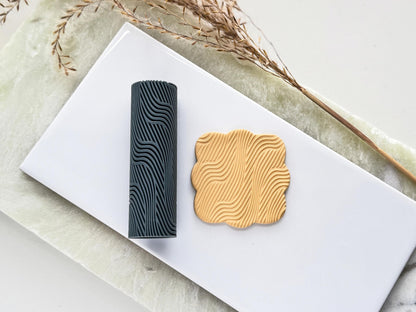 Grey texture roller with wavy line pattern for leaving embossed textures in clay. Next to it is a beige polymer clay cut out sample.