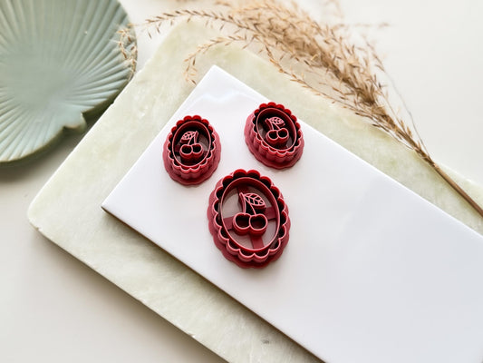 Vintage style cherry frame shaped clay cutters. Features scalloped edge, inner oval, double cherry and leaves stamp details. The image shows a small mirrored pair of cutters and a single large cutter.