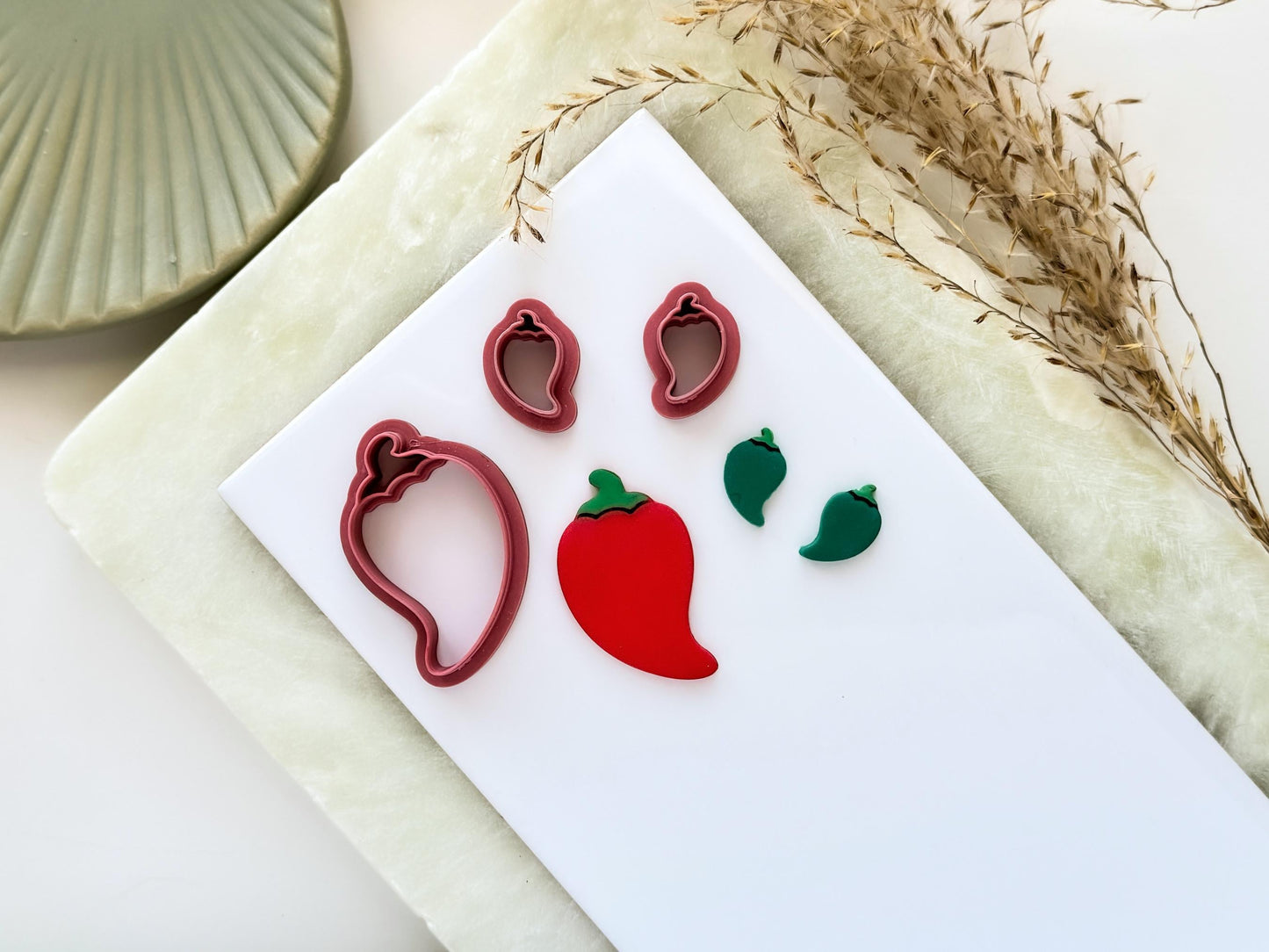 Short chilli pepper shaped clay cutters, the image shows a small mirrored pair and a larger single cutter. Next to them are polymer clay cut out samples in red and green on a white tile.