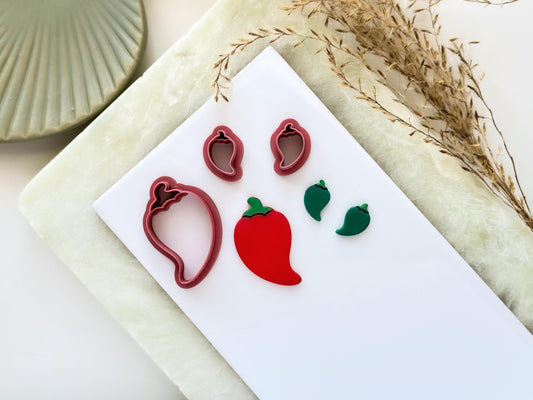 Short chilli pepper shaped clay cutters, the image shows a small mirrored pair and a larger single cutter. Next to them are polymer clay cut out samples in red and green on a white tile.