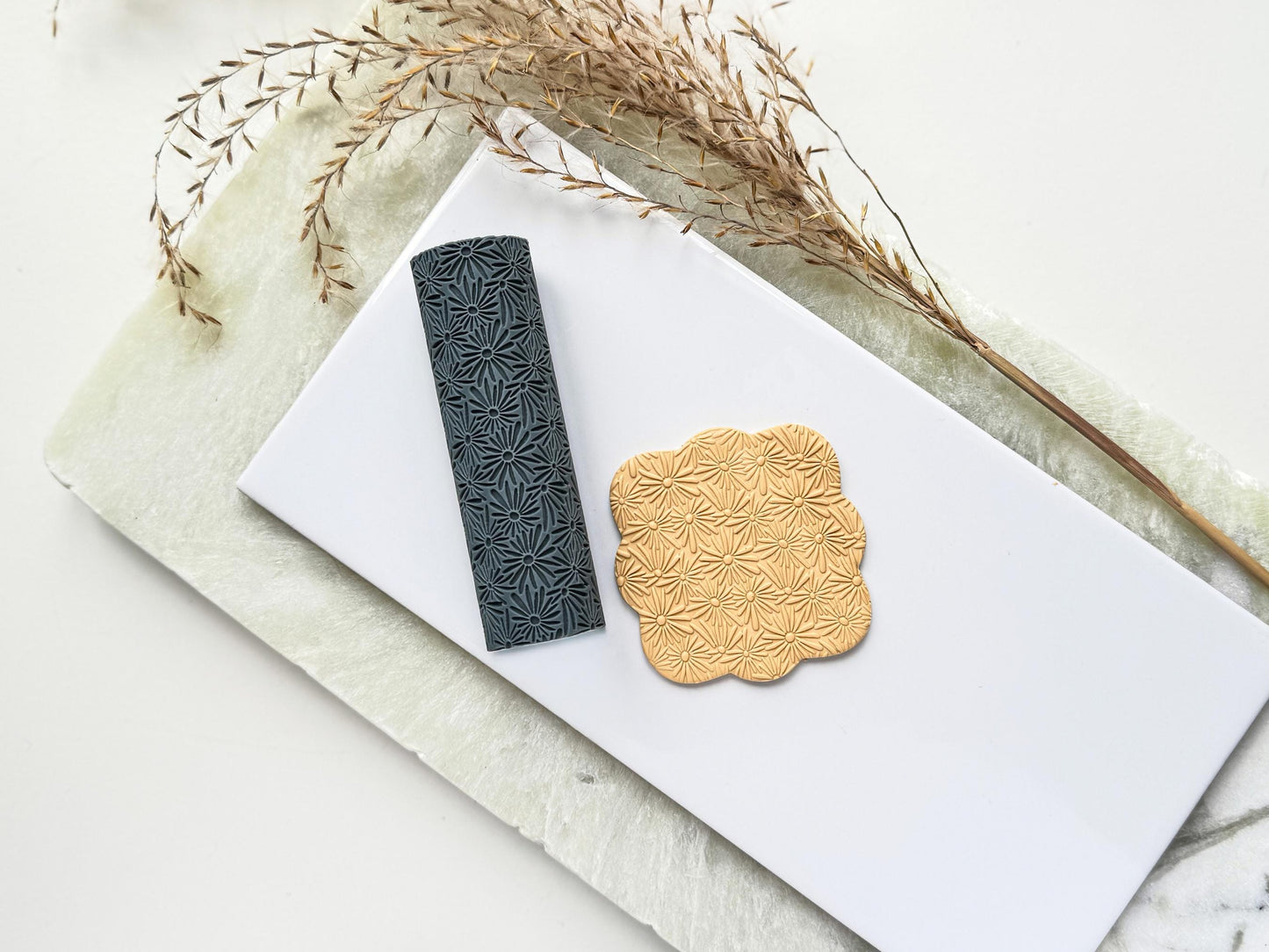 A texturising tool for use on clay, it features a seamless thin petal daisy flower pattern. Next to the clay tool is a square polymer clay cut out with the floral embossed pattern.