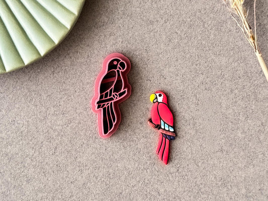 Parrot shaped clay cutter in dark pink. The parrot is sat on a tree branch and features wing, beak and tail feather details. next to the cutter is a polymer clay cut out sample painted in tropical colours.