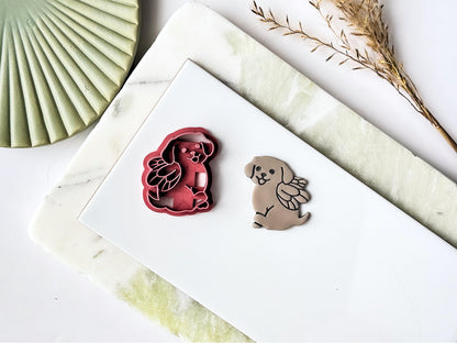 Pink sitting fairy dog shaped clay cutter with wings detail, next to the cutter is a polymer clay cut out sample earring. Both are placed on a white tile with a green marble background.