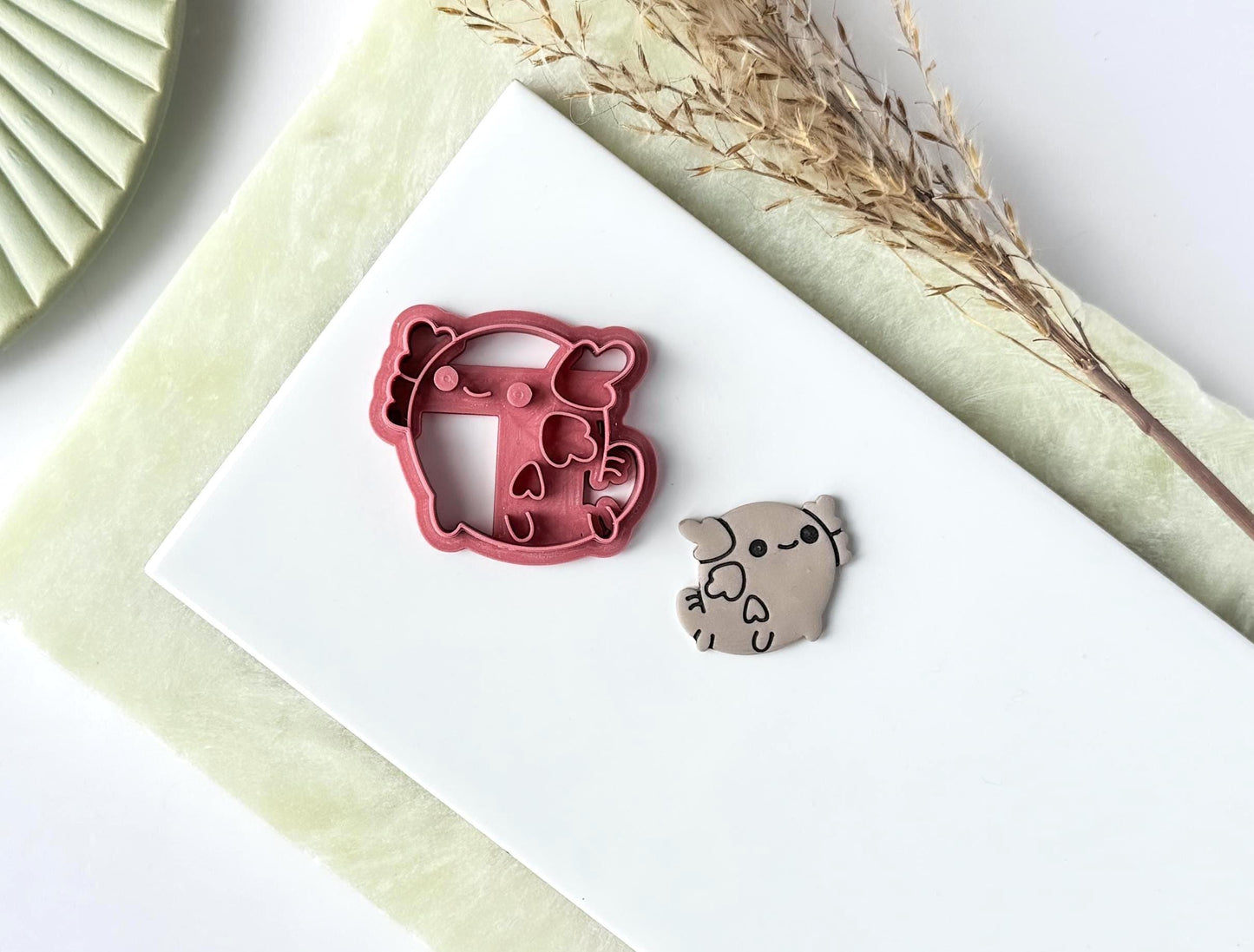 Cartoon style axolotl clay cutter in dark pink with cute smiling face detail. Next to it is a polymer clay cut out sample with the stamp details painted in dark grey. Both are on a tile.