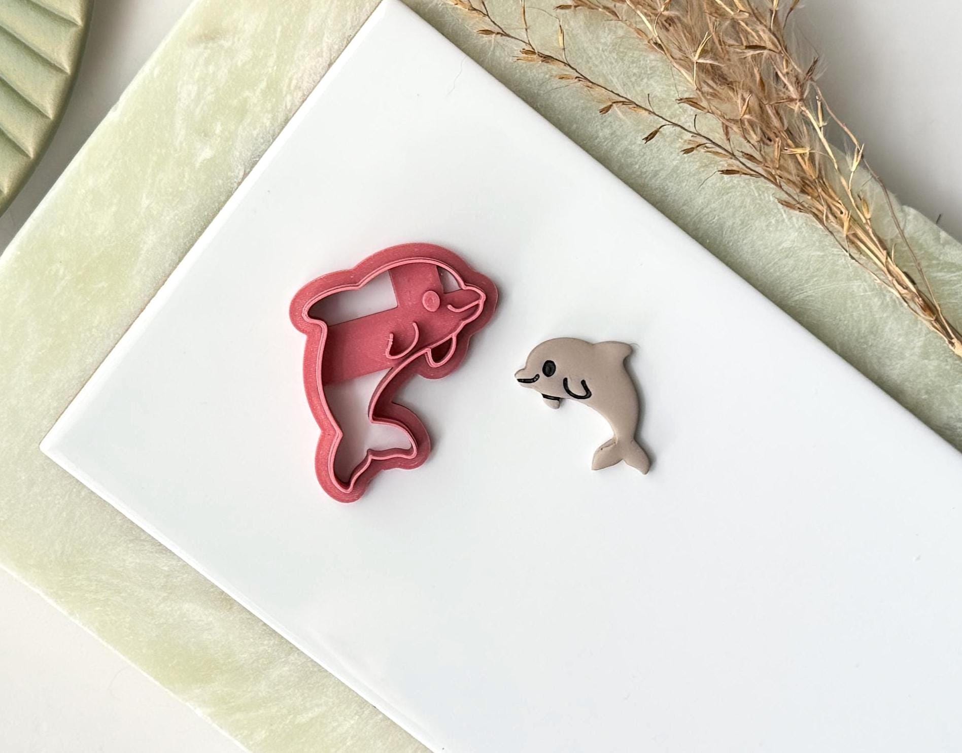 Pink plastic craft cutter in the shape of a cute cartoon dolphin with fins and smiling face. Next to the cutter is a polymr clay cut out sample. Both are on a white tile.