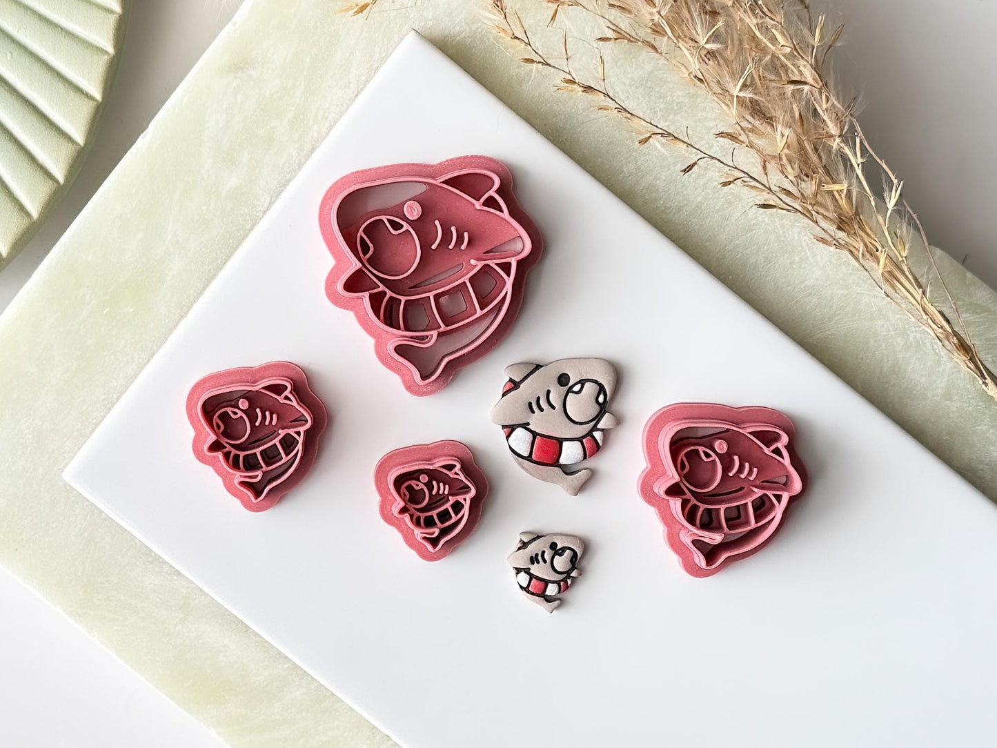 Various sizes of shark clay cutter, from 1.5cm to 4.5cm. There are painted polymer clay cut outs next to them.
