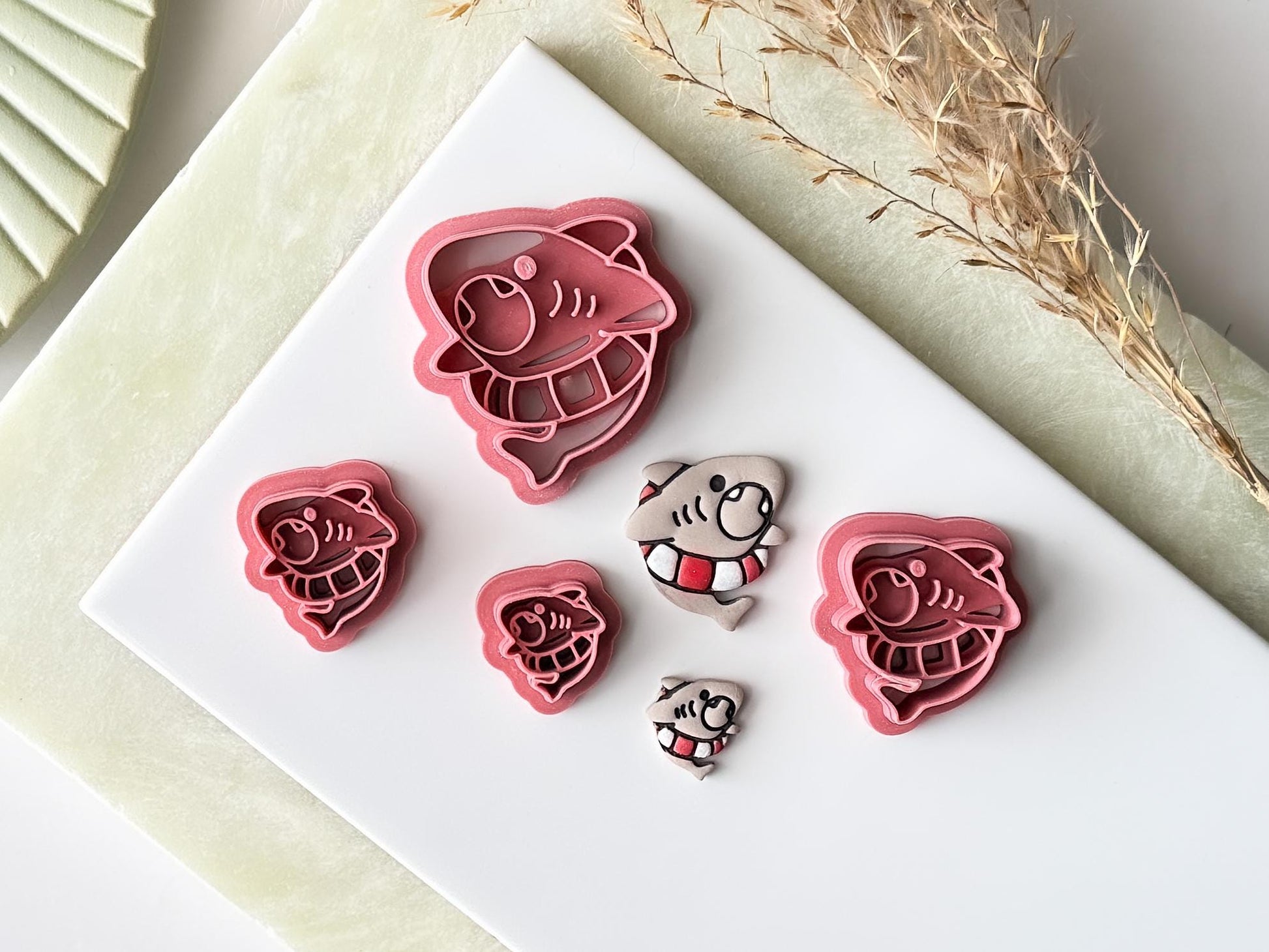 Various sizes of shark clay cutter, from 1.5cm to 4.5cm. There are painted polymer clay cut outs next to them.