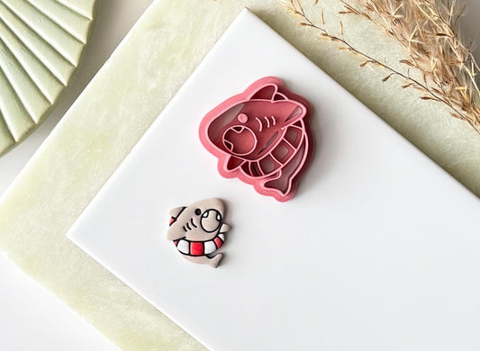 Cute shark stuck in a life ring shaped clay cutter with mouth, fin and gill details. Next to the cutter is a polymer clay cut out sample, painted. The cutter is for making ocean themed clay jewellery and cake decorations.