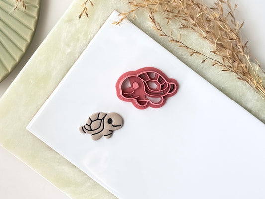 Cute cartoon style sea turtle shaped clay cutter with shell and face detail. Next to the cutter is a taupe coloured polymer clay turtle made from the cutter. Both are on a clay baking tile.