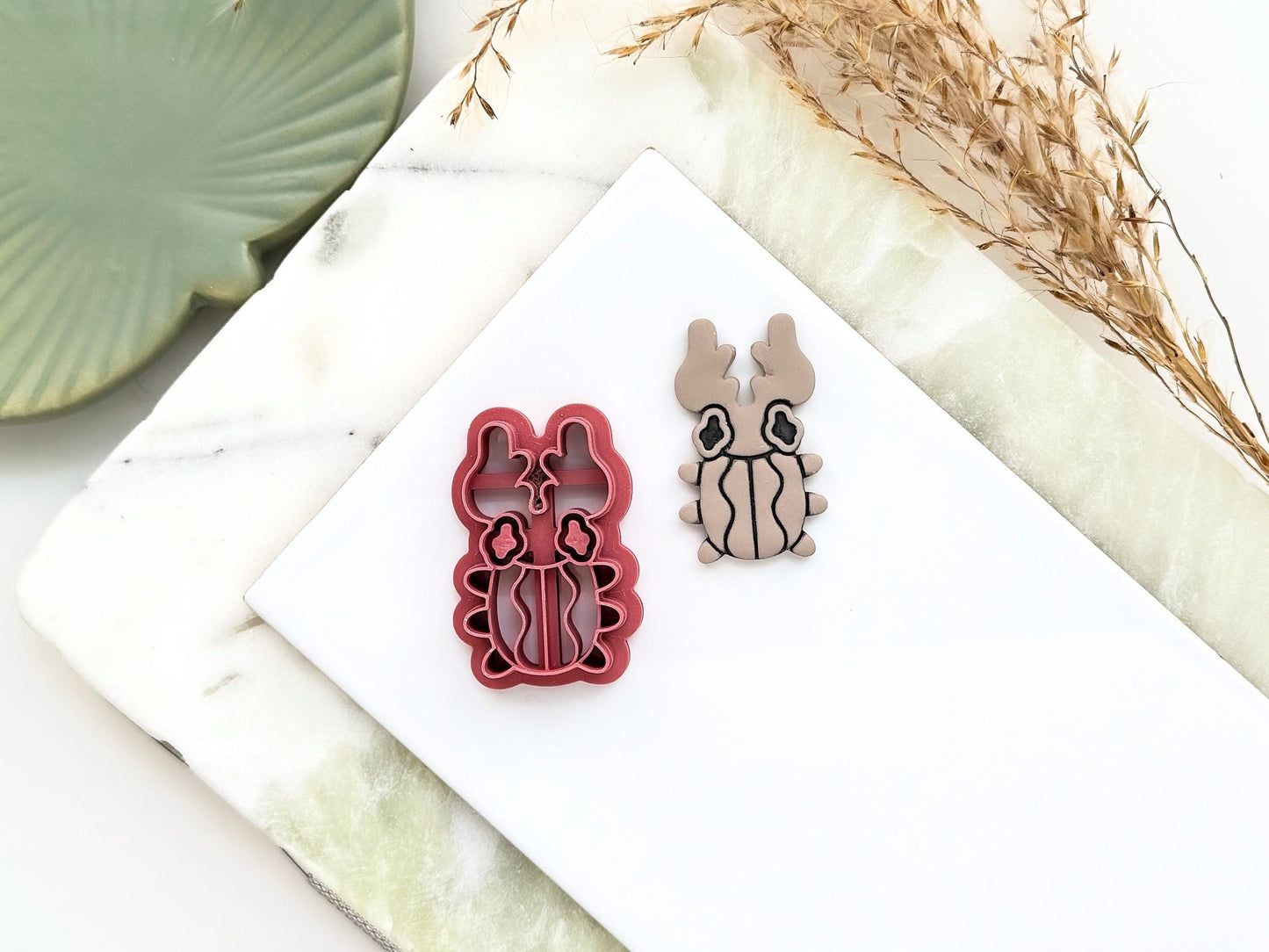 Horn beetle shaped clay cutter with wavy shell, eyes and large horns. Next to the cutter is a polymr clay cut out sample earring