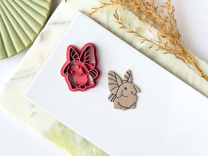 Pink cute rabbit shaped clay cutter with fairy wings detail, next to the cutter is a polymer clay cut out sample earring. Both are placed on a white tile with a green marble background.