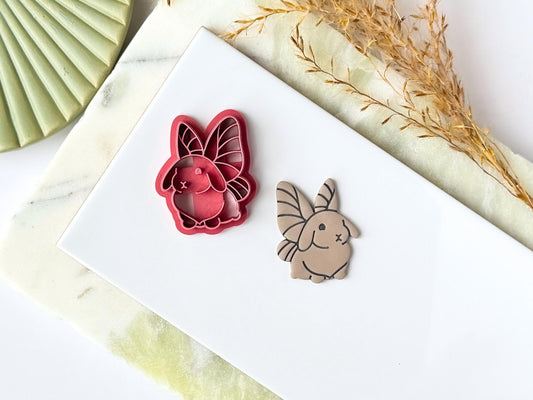 Pink cute rabbit shaped clay cutter with fairy wings detail, next to the cutter is a polymer clay cut out sample earring. Both are placed on a white tile with a green marble background.