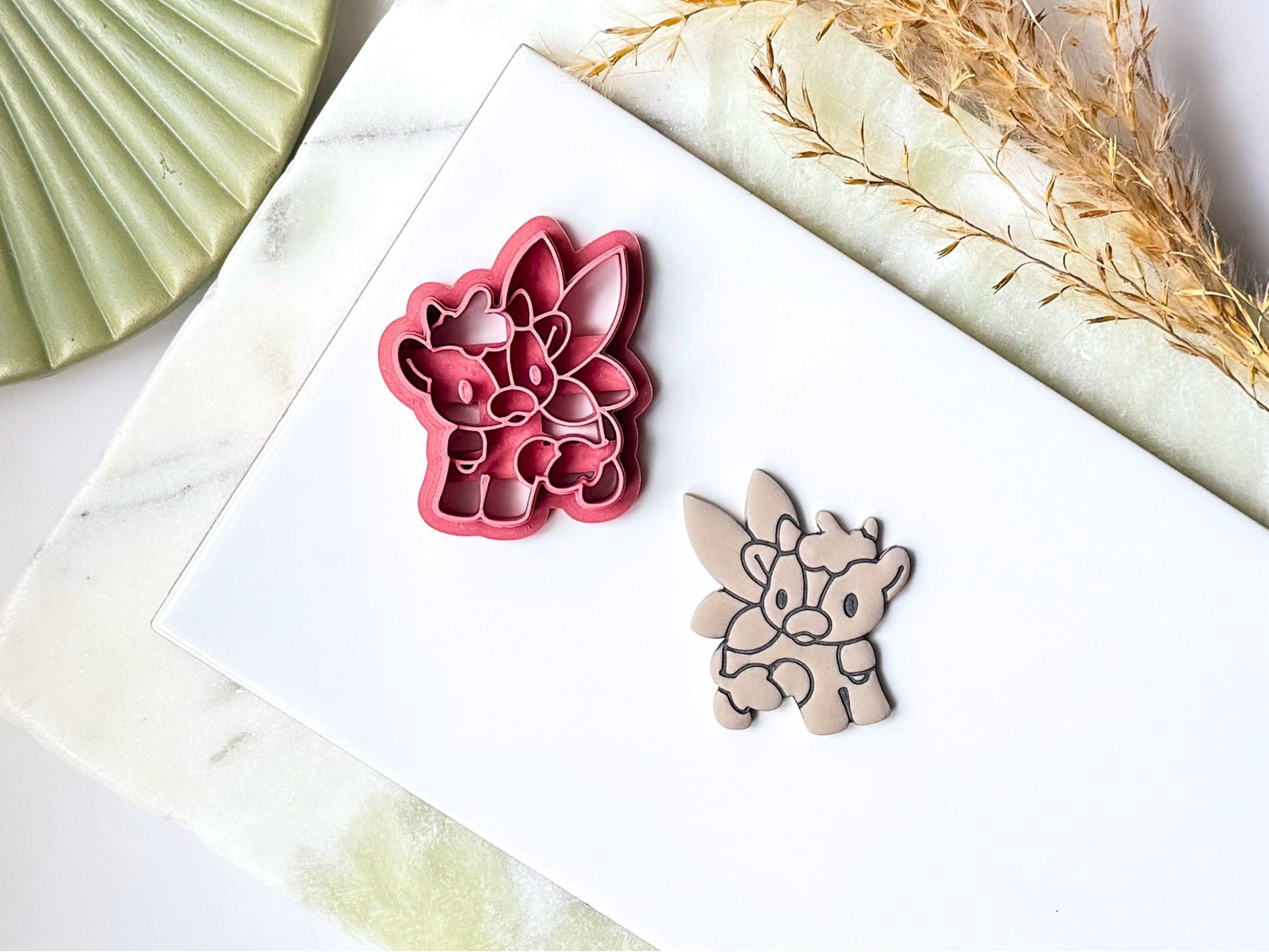 Pink cute cow shaped clay cutter with fairy wings, horns, spots and bell stamp details, next to the cutter is a polymer clay cut out sample earring. Both are placed on a white tile with a green marble background.