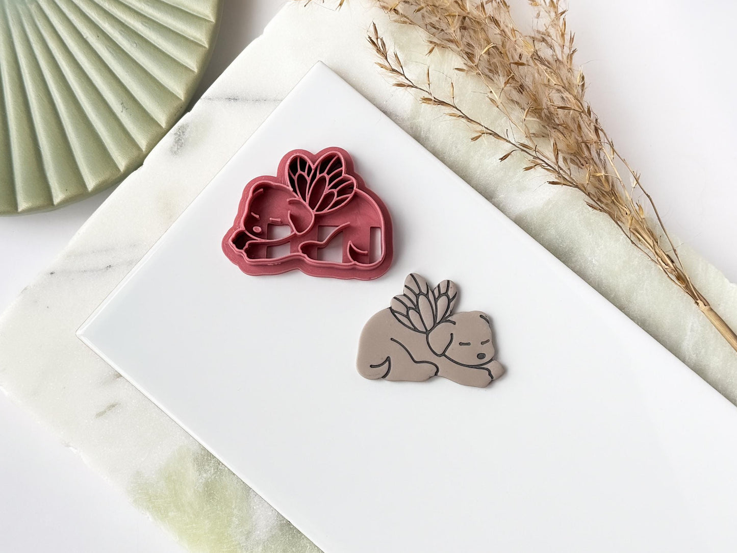 A pink polymer clay cutter in the shape of a sleeping dog with fairy wings next to a clay cut out sample. Both items are sat on a white tile