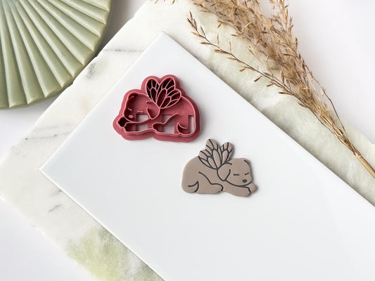 A pink polymer clay cutter in the shape of a sleeping dog with fairy wings next to a clay cut out sample. Both items are sat on a white tile