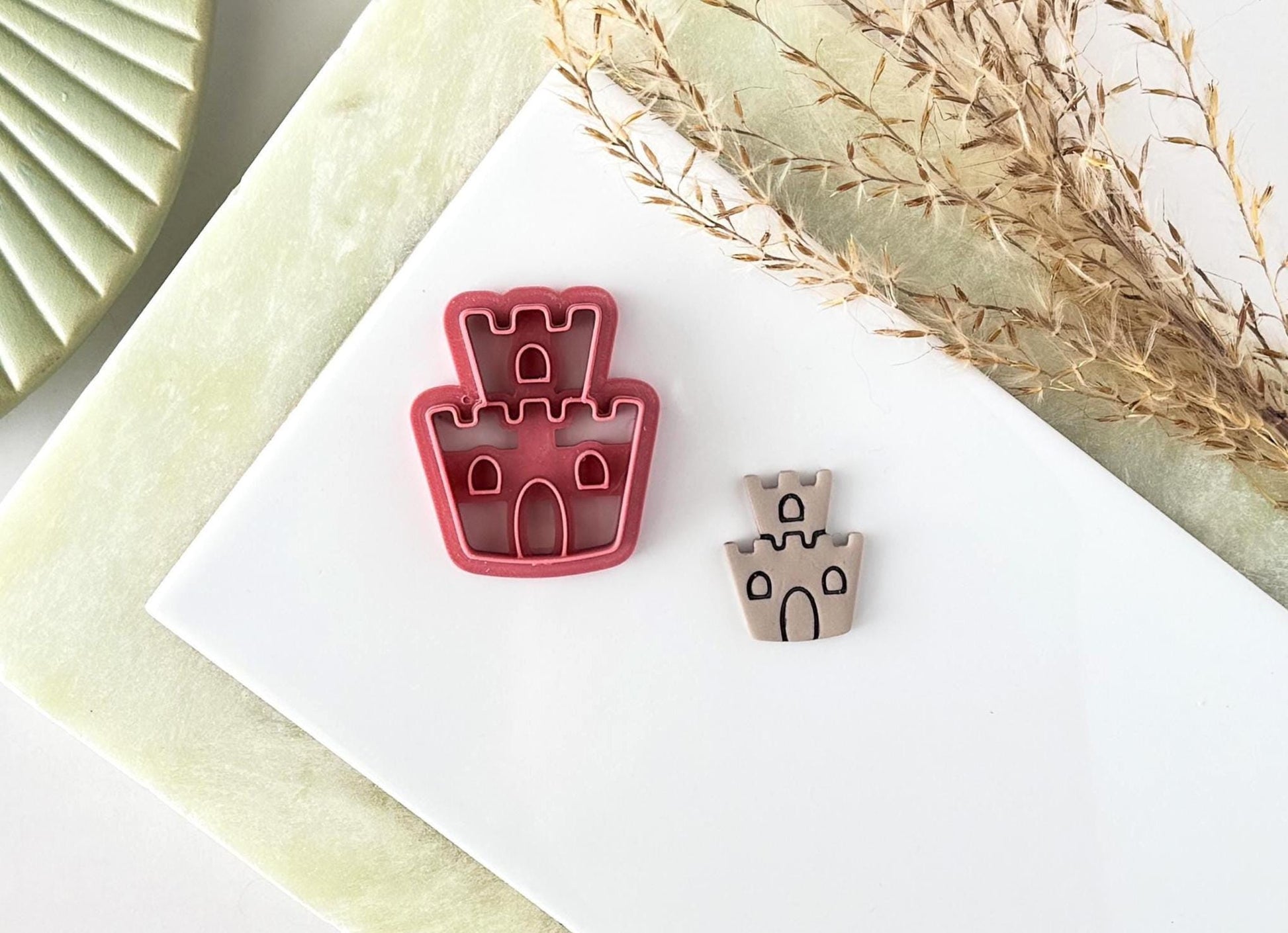 Pink plastic sandcastle-shaped craft cutter with castle style top, windows and door detail. Next to the cutter is a polymer clay cut out sample with the imprint details painted dark gray.