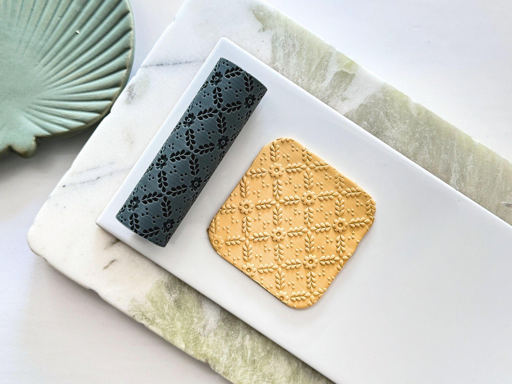 Floral lattice patterned texture roller for making detailed impressions in slabs on clay. Next to the rolling pin is a polymer clay square with the pattern embossed into the surface. The pattern is a criss-cross style featuring leaves and simple style flower as the main texture and small clusters of dots in the spaces. Both items are on marble green and white tiles.