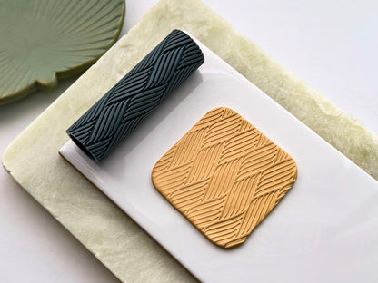 Grey texture roller with rattan weave style geometric line pattern for leaving embossed textures in clay. Next to it is a beige polymer clay cut out sample.