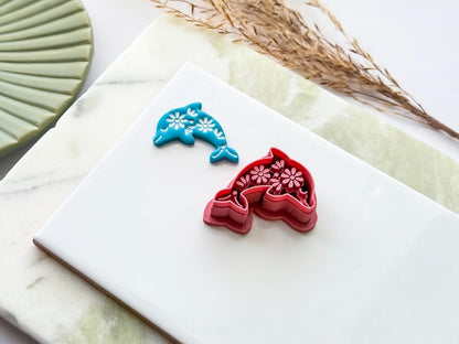 Side view of a dolphin-shaped polymer clay cutter with decorative floral embossing shown alongside a blue clay impression. UK-made crafting tool for polymer clay, fondant, jewellery making and creative craft projects.