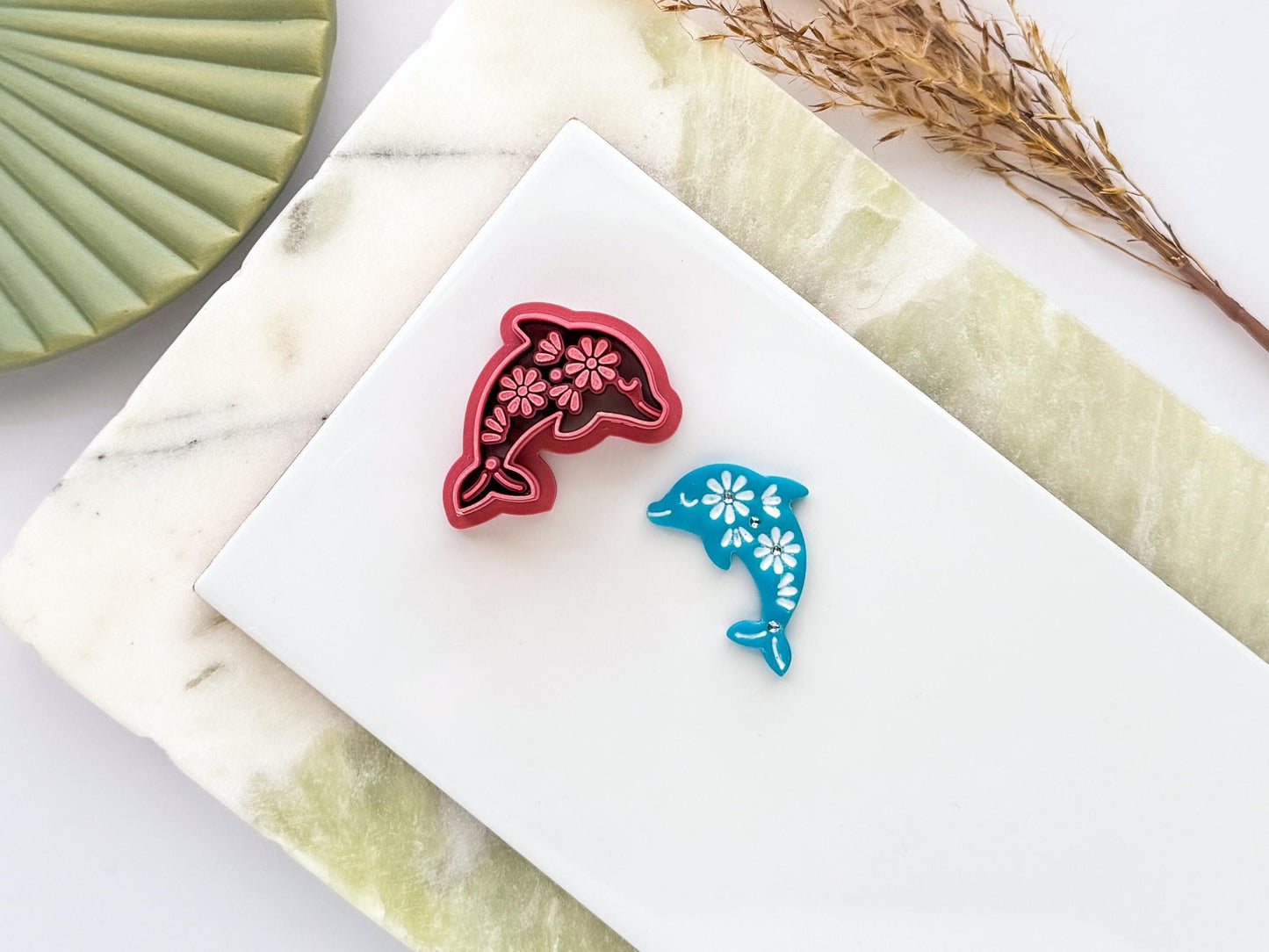 Floral dolphin polymer clay cutter in deep red PLA displayed beside a matching blue clay cut-out. The dolphin design features intricate flower details and is photographed on a white ceramic tile with a marble-effect background.