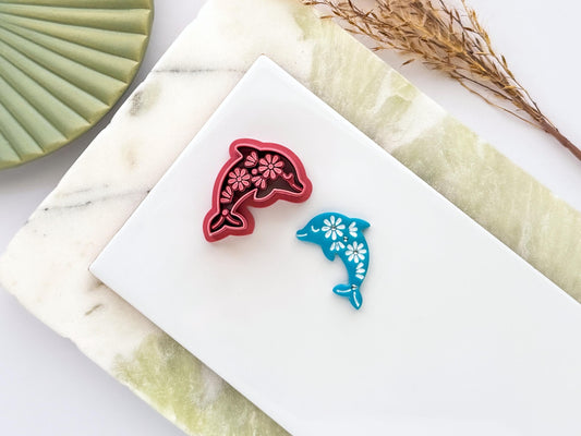 Floral dolphin polymer clay cutter in deep red PLA displayed beside a matching blue clay cut-out. The dolphin design features intricate flower details and is photographed on a white ceramic tile with a marble-effect background.