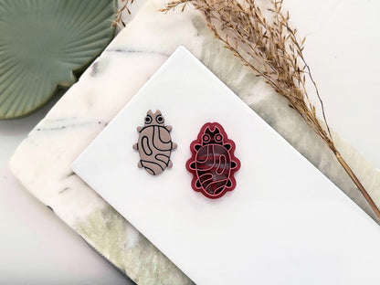 Abstract style leaf bug-shaped clay cutter in pink with embossing details. Next to the cutter is a polymer clay cut out made with the tool.