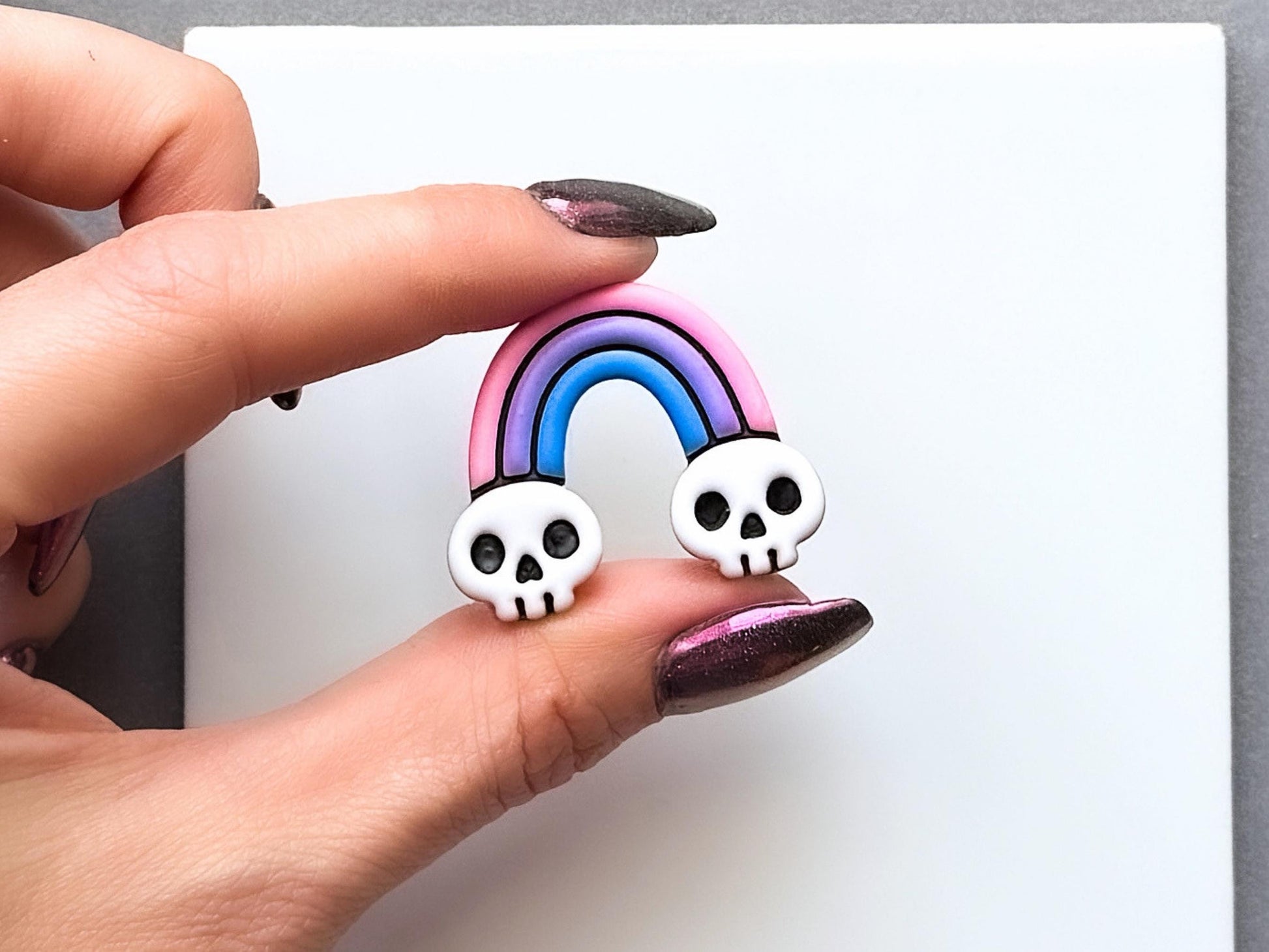 Finished summerween polymer clay rainbow with skull details at each end, held between fingers to show size. The gothic rainbow design features pink, purple and blue arches with white skull accents