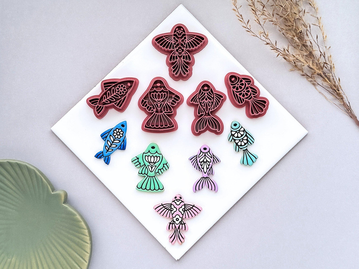 Collection of five ornamental fish polymer clay cutters displayed with matching colourful clay cut-outs. The set includes detailed folk art-inspired fish designs photographed on a white ceramic tile.