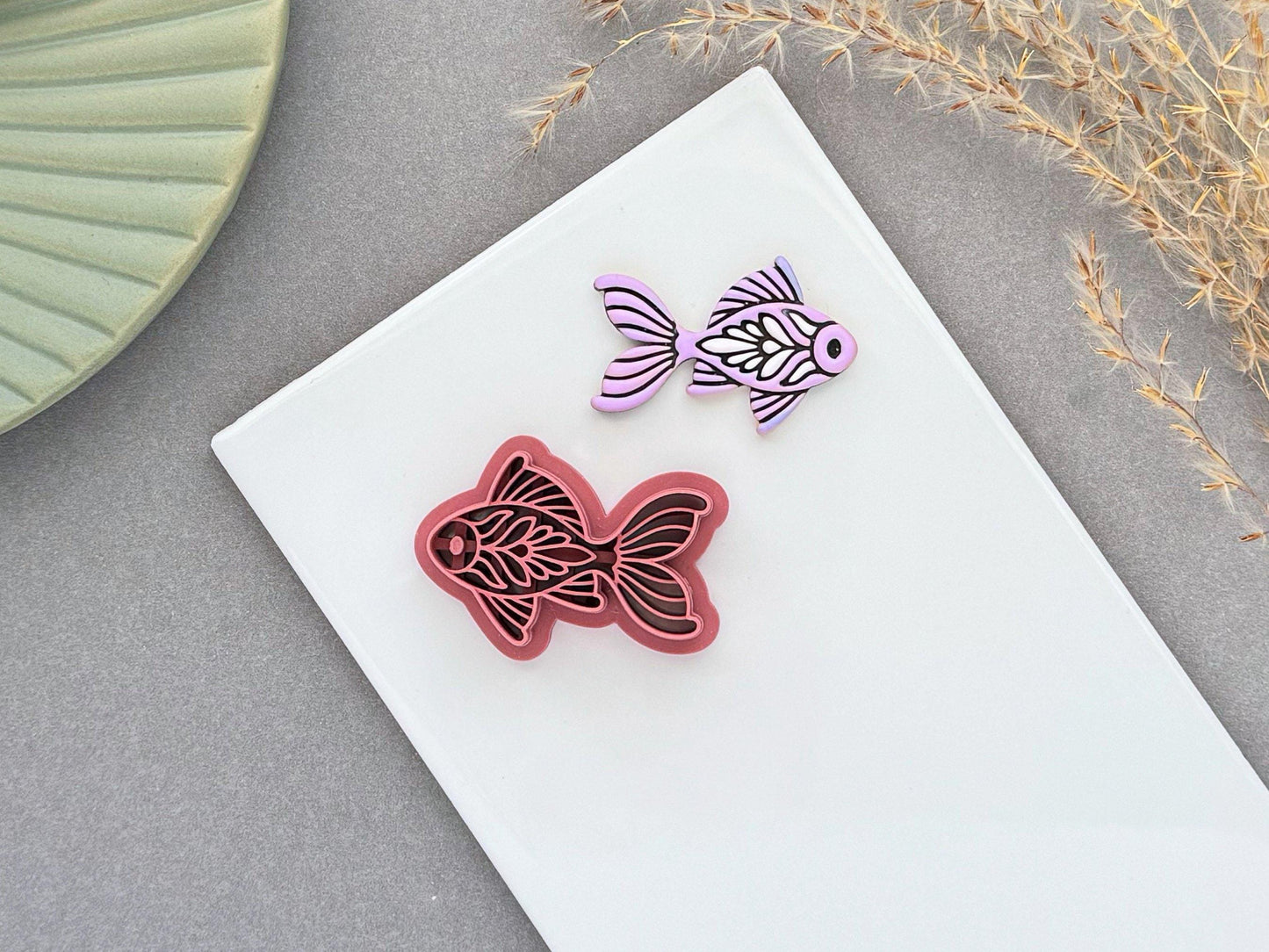 Ornamental fish polymer clay cutter displayed beside a matching lavender clay cut-out. The folk art-inspired fish design features flowing fins and decorative leaf details and is photographed on a white ceramic tile.