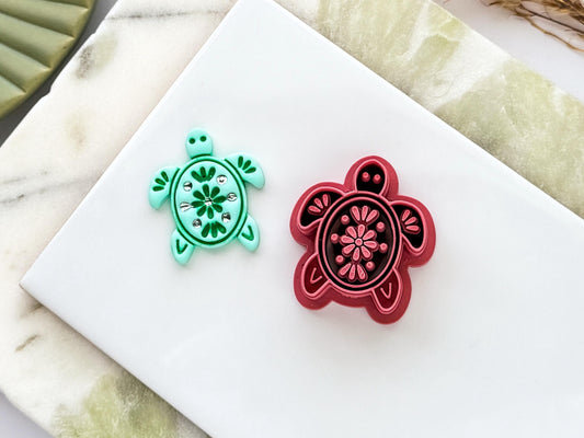 Floral sea turtle polymer clay cutter in deep red PLA shown alongside a matching mint green clay cut-out. The embossed turtle design features intricate floral details and is displayed on a white ceramic tile with a marble-effect background.