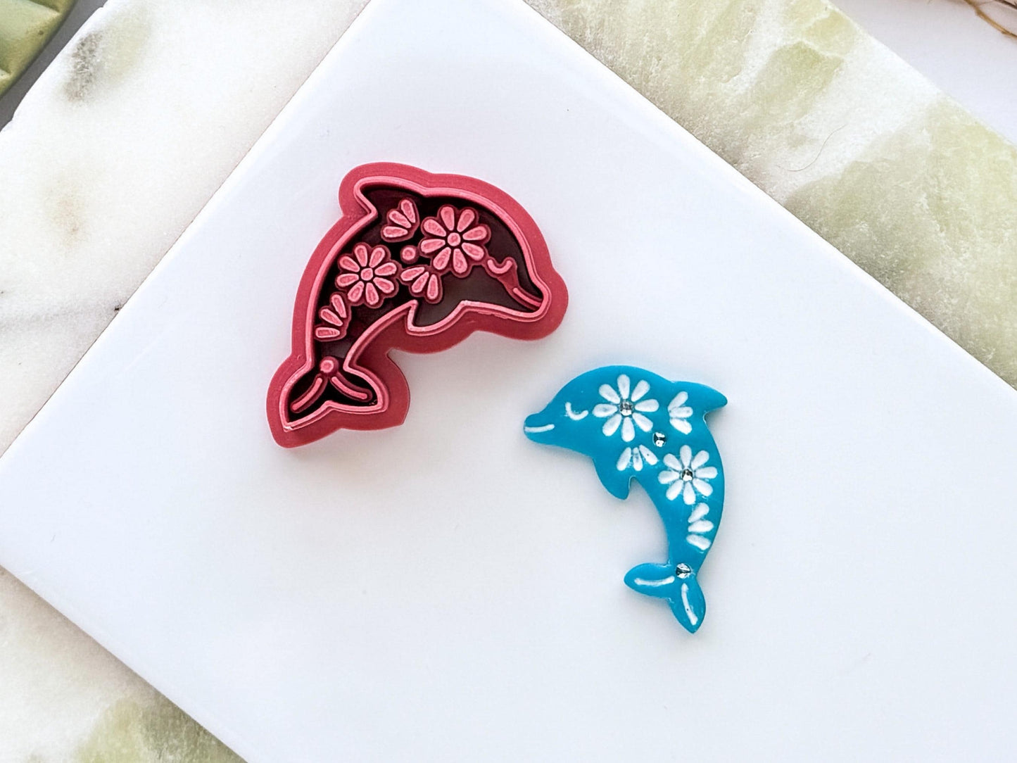 Close up of floral fok art dolphin polymer clay cutter in dark pink PLA displayed beside a matching blue clay cut-out. The dolphin design features intricate flower details and is photographed on a white ceramic tile with a marble-effect background.