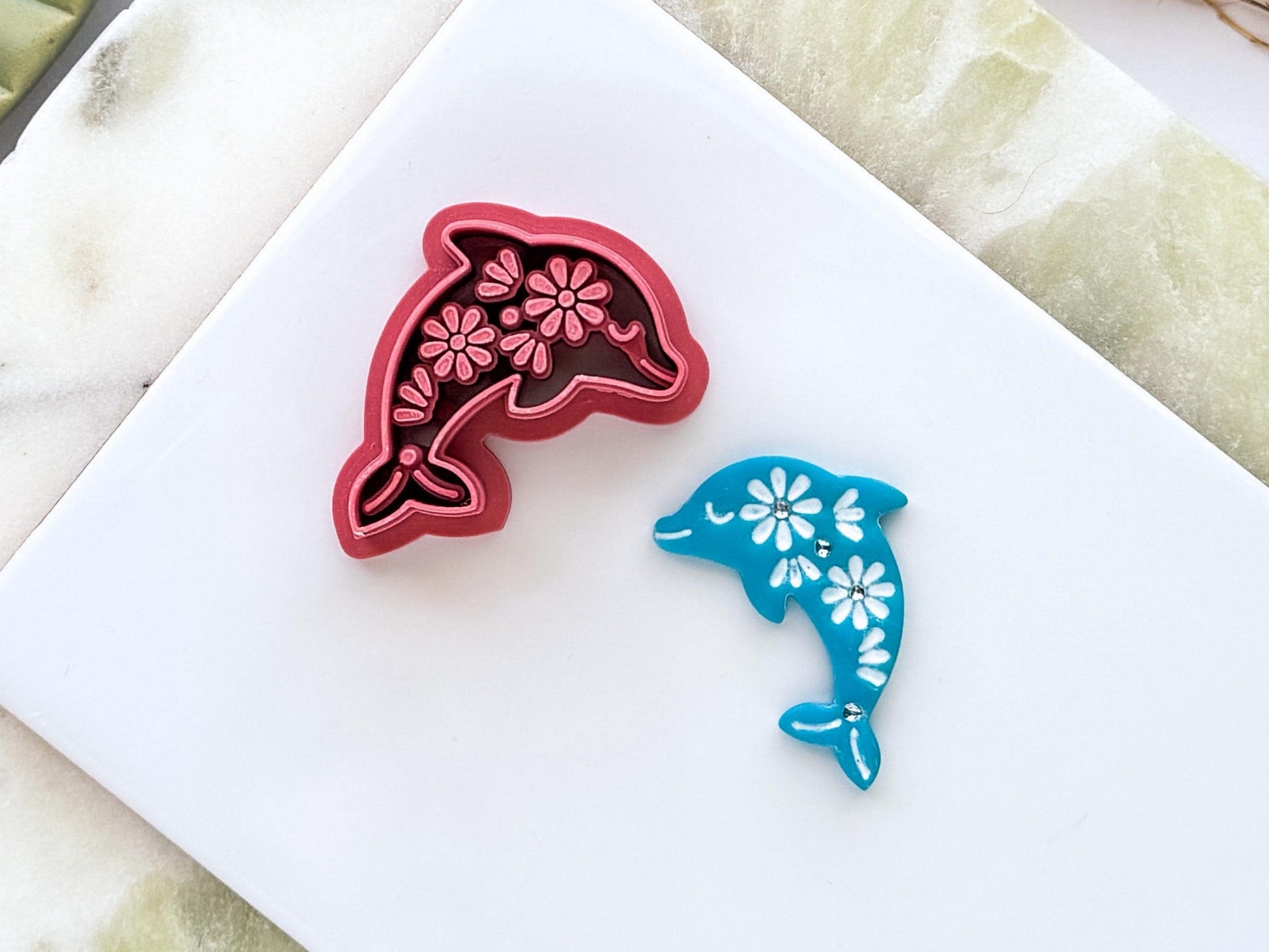 Close up of floral fok art dolphin polymer clay cutter in dark pink PLA displayed beside a matching blue clay cut-out. The dolphin design features intricate flower details and is photographed on a white ceramic tile with a marble-effect background.