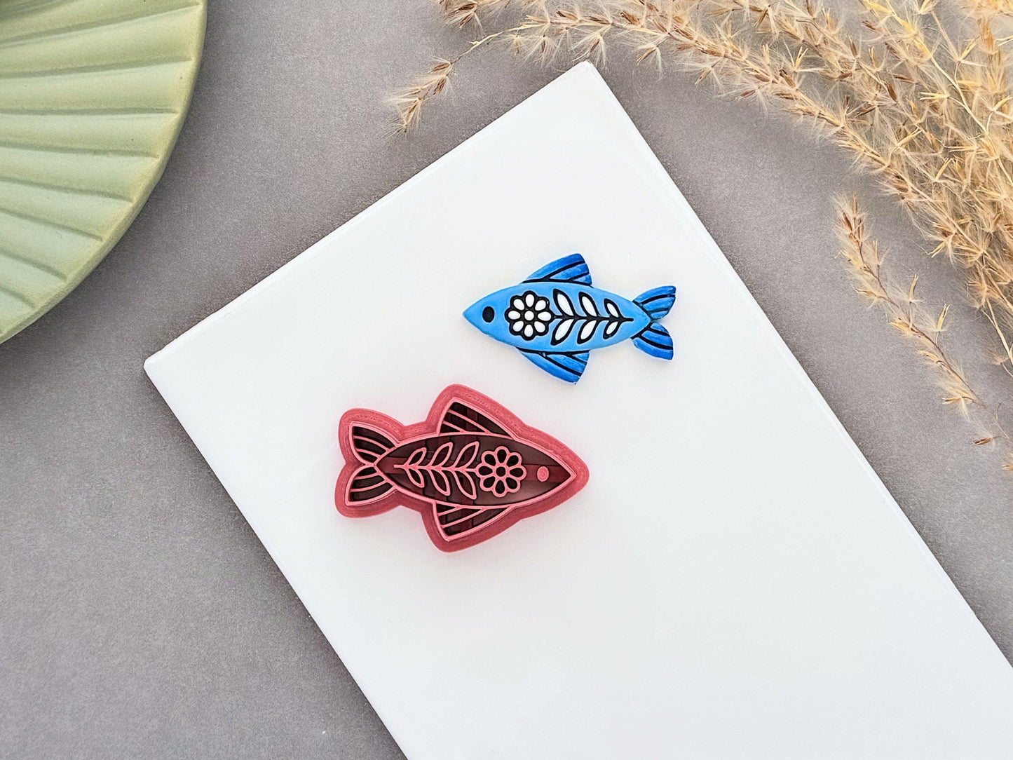 Folk art fish polymer clay cutter displayed beside a matching blue clay cut-out. The ornamental fish design features floral and leaf details and is photographed on a white ceramic tile.