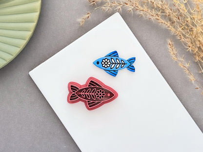 Folk art fish polymer clay cutter displayed beside a matching blue clay cut-out. The ornamental fish design features floral and leaf details and is photographed on a white ceramic tile.