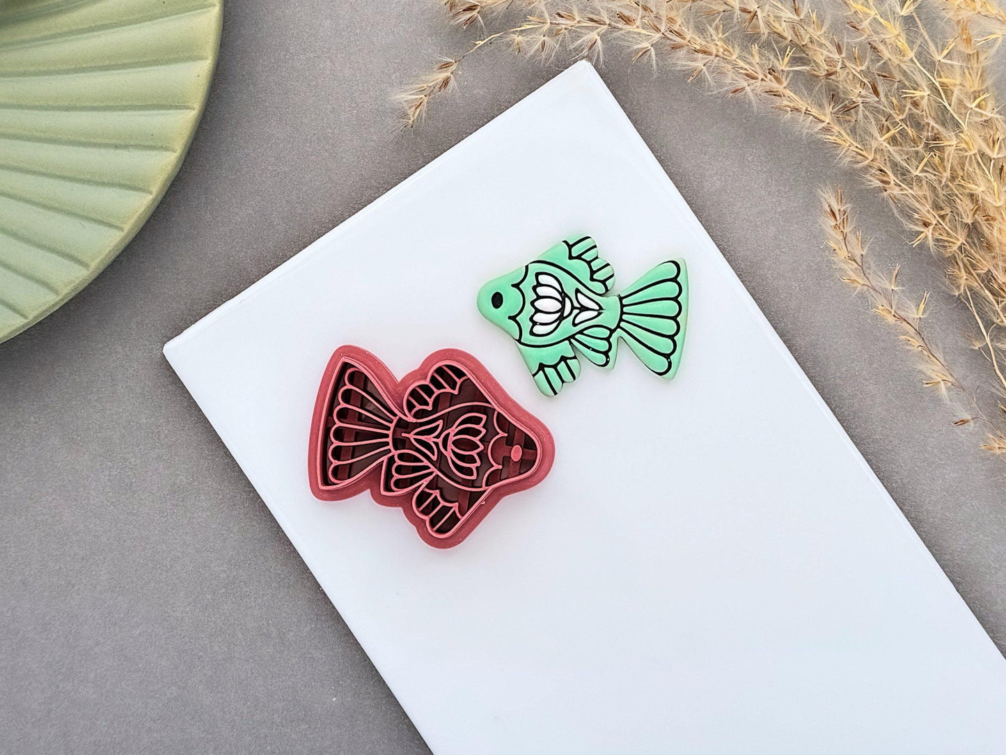 Folk art fish polymer clay cutter with detailed floral and fin designs, shown next to a mint green clay cut-out. UK-made clay tool for earrings, jewellery making, fondant decorating and handmade craft projects.