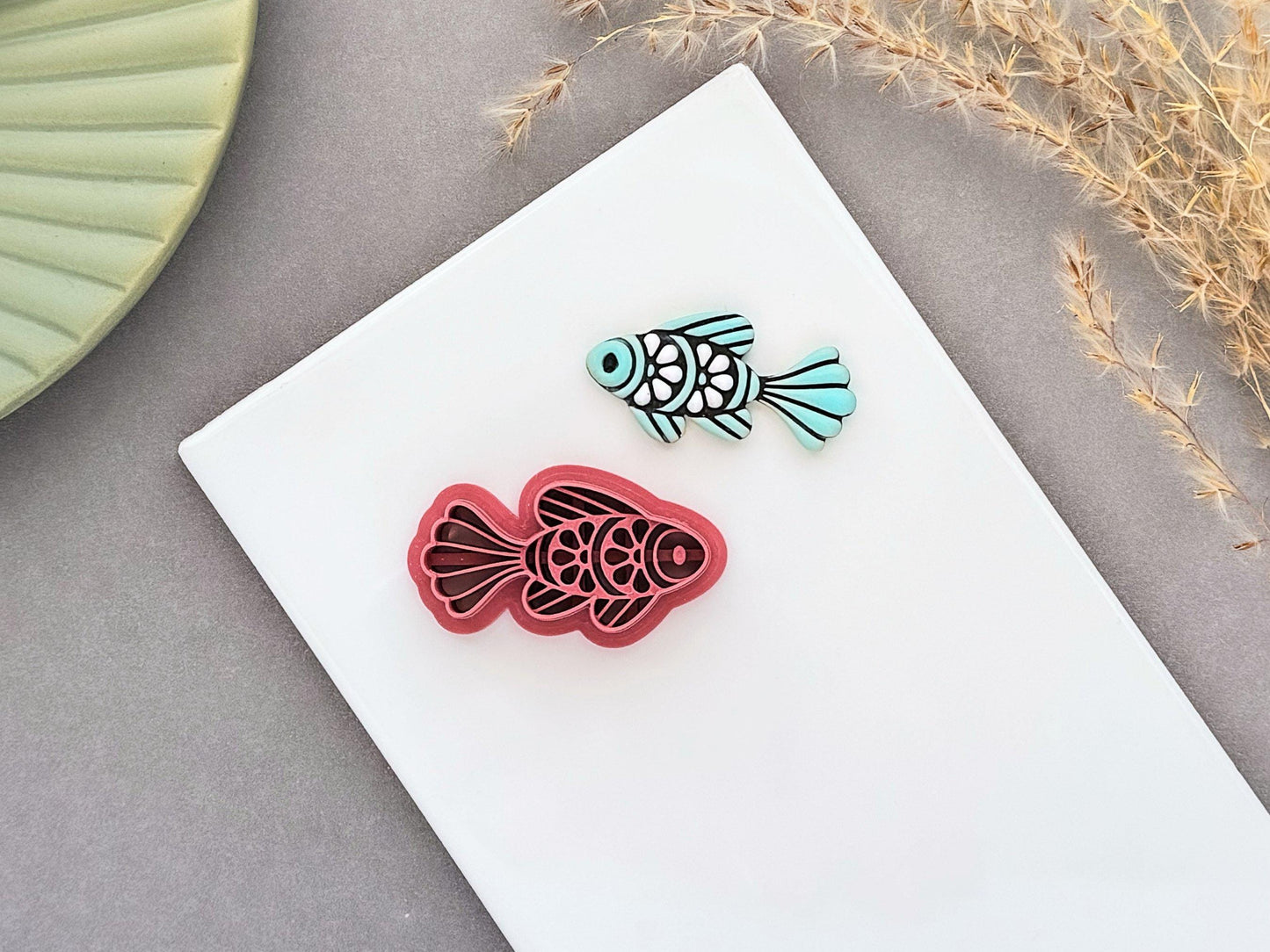 Ornamental fish polymer clay cutter displayed beside a matching pale teal clay cut-out. The folk art-inspired fish design features decorative scale and fin details and is photographed on a white ceramic tile.