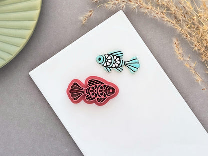 Ornamental fish polymer clay cutter displayed beside a matching pale teal clay cut-out. The folk art-inspired fish design features decorative scale and fin details and is photographed on a white ceramic tile.