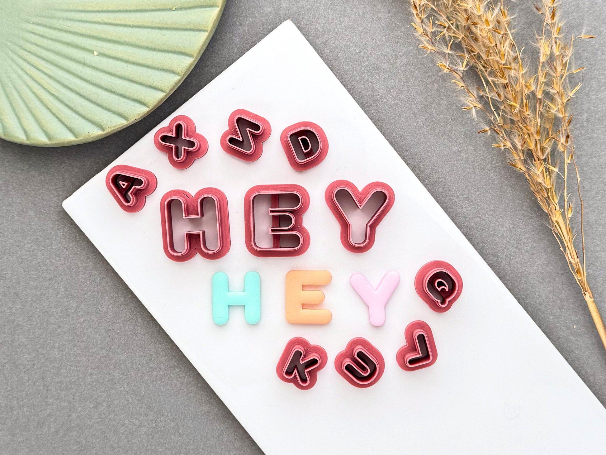 Uppercase alphabet polymer clay letter cutters with matching pastel clay letters spelling "HEY" and coordinating lowercase letter cutters displayed on a white tile.