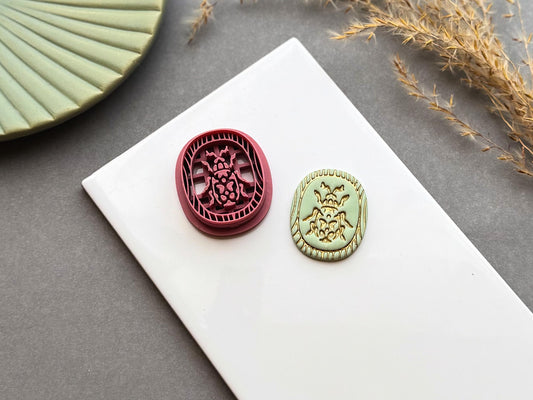 Decorative beetle polymer clay cutter displayed beside a matching finished polymer clay embellishment featuring an ornate beetle design within a decorative Egyptian-inspired border.