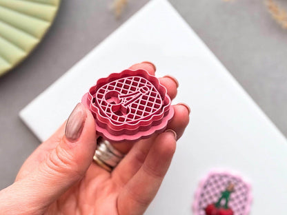 Cherry polymer clay cutter held in a hand for scale, showcasing the detailed cherry design, decorative lattice background, scalloped frame and fine cutting edge.
